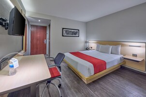 Habitación de hotel con cama, mesitas de noche, escritorio con silla, televisor montado en la pared y una puerta.