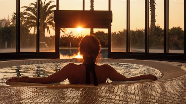 A person in a hot tub with a view of palm trees and a sunset through large windows.