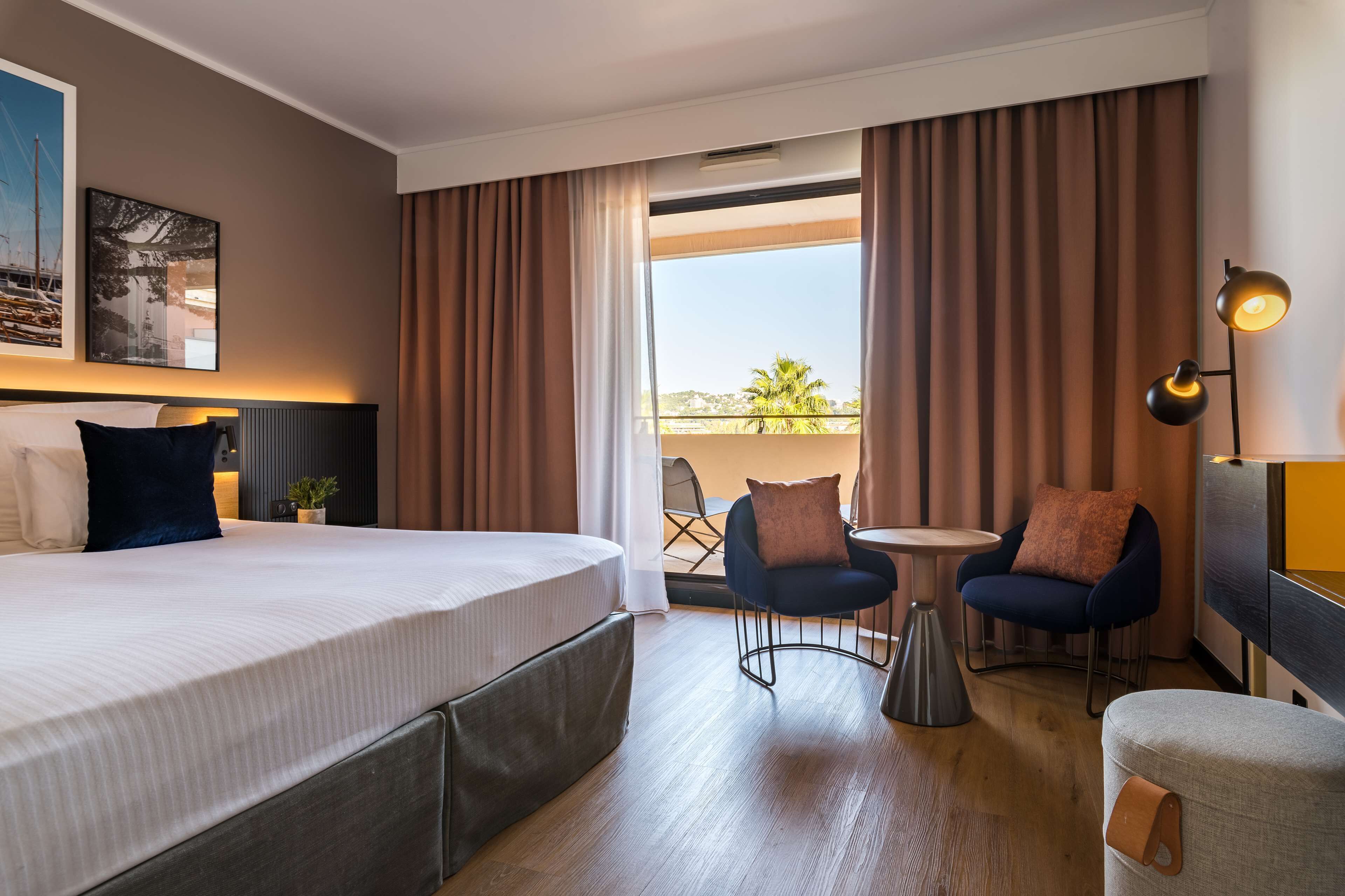 A hotel room with a large bed, a small table, two chairs, and a balcony with curtains.