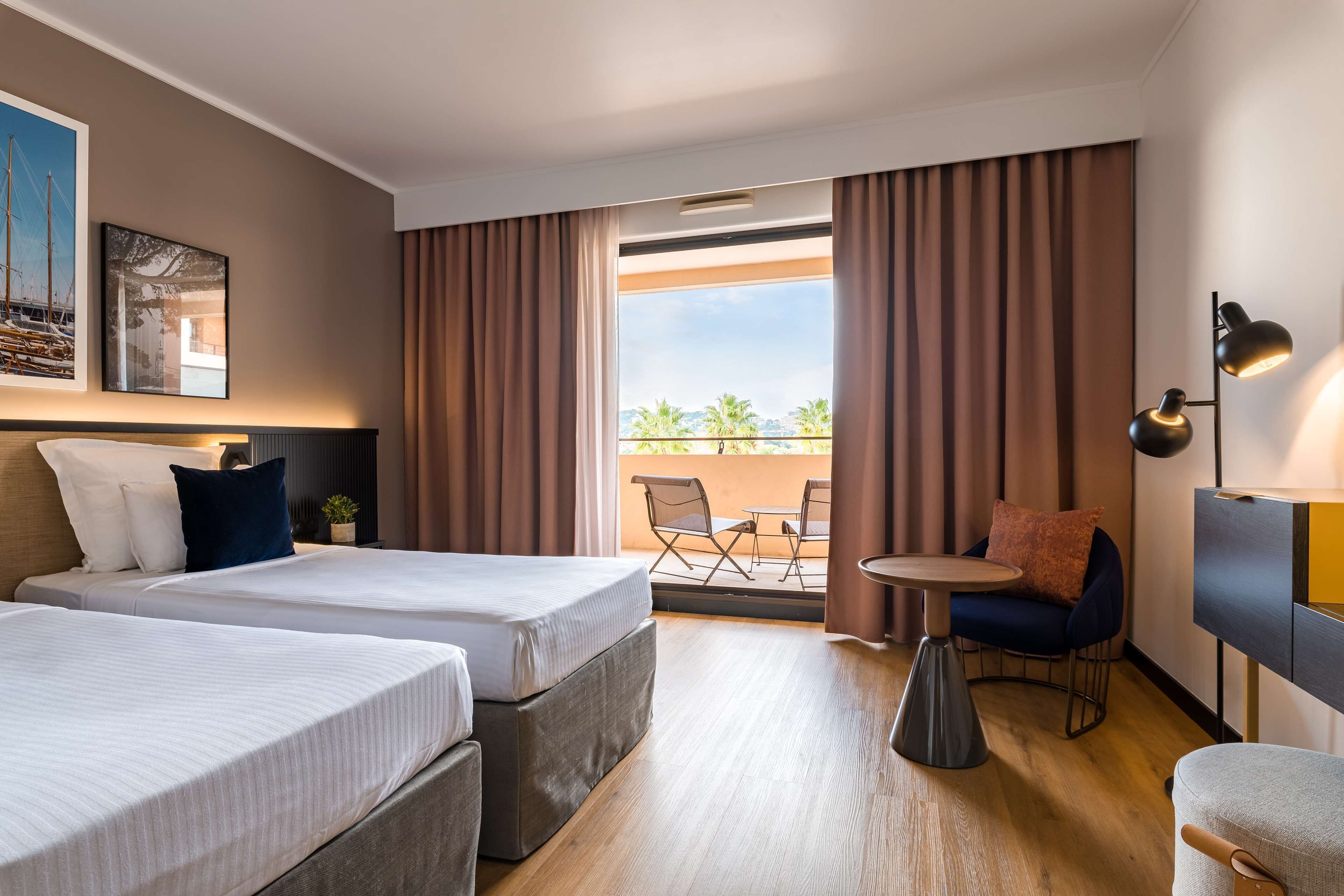 Een hotelkamer met twee bedden, een klein balkon met een tafel en stoelen, en uitzicht op palmbomen.
