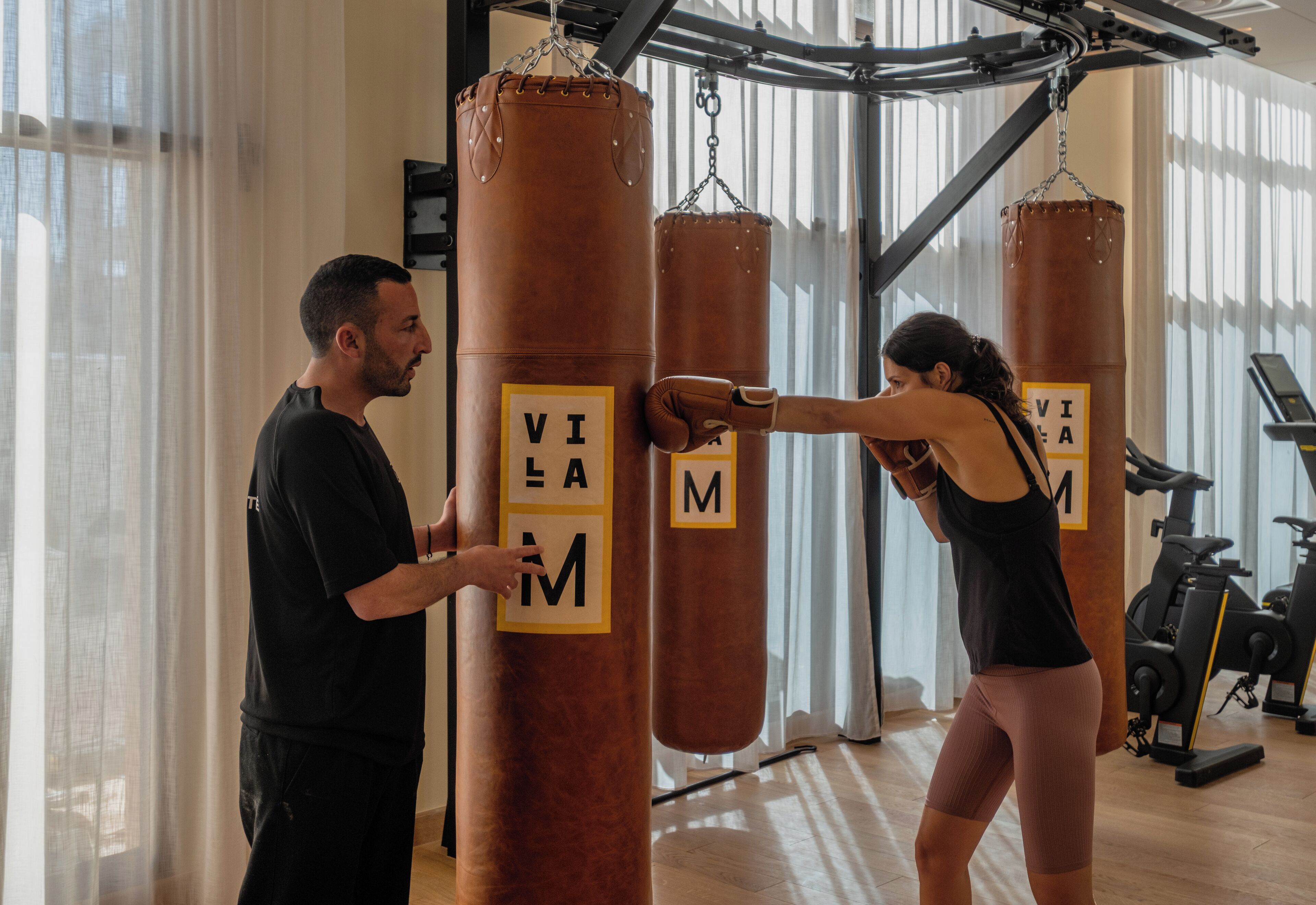 Two people training with punching bags in a gym with exercise equipment in the background.