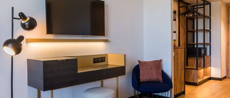A modern hotel room with a flat-screen TV mounted on the wall, a desk with a lamp, a chair, and a built-in wardrobe with shelves.