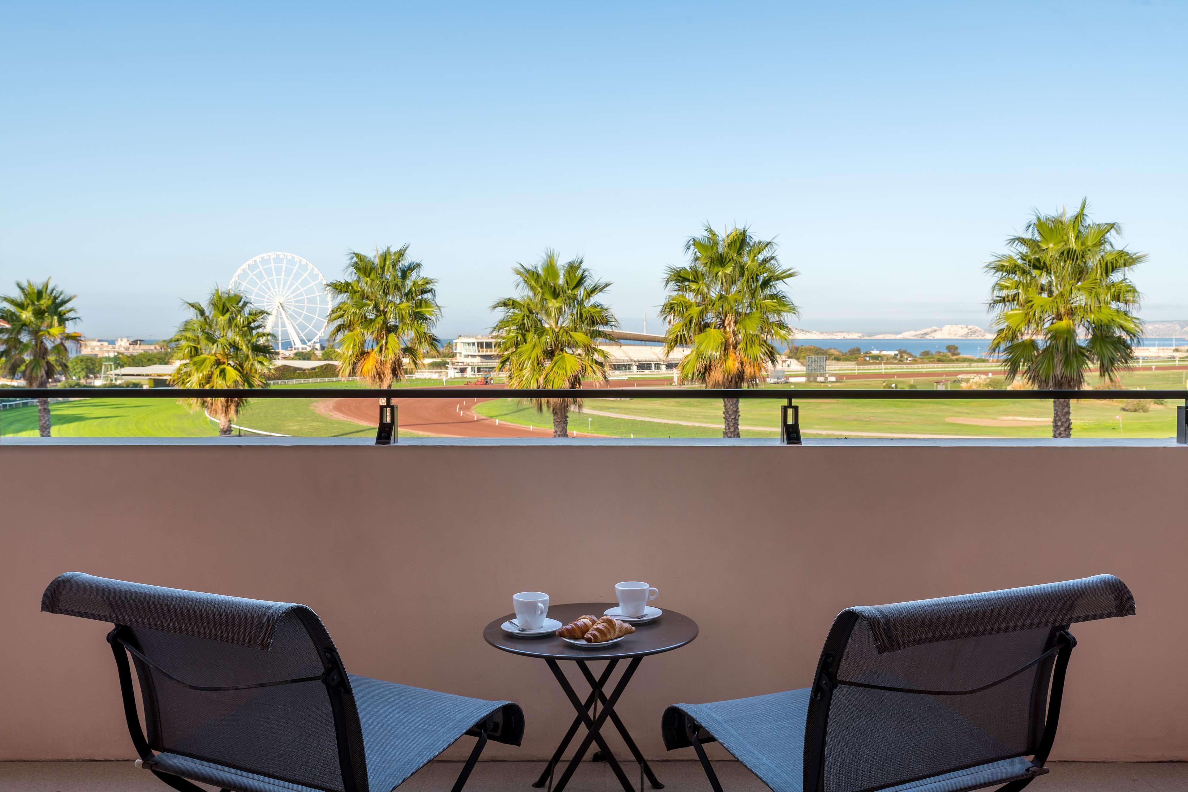 A balcony with two chairs and a small table with cups and croissants, overlooking a baseball field and a Ferris wheel.