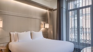 Una cama grande con ropa de cama blanca, una mesita de noche, una lámpara de pared y vistas a un balcón a través de cortinas de gasa.
