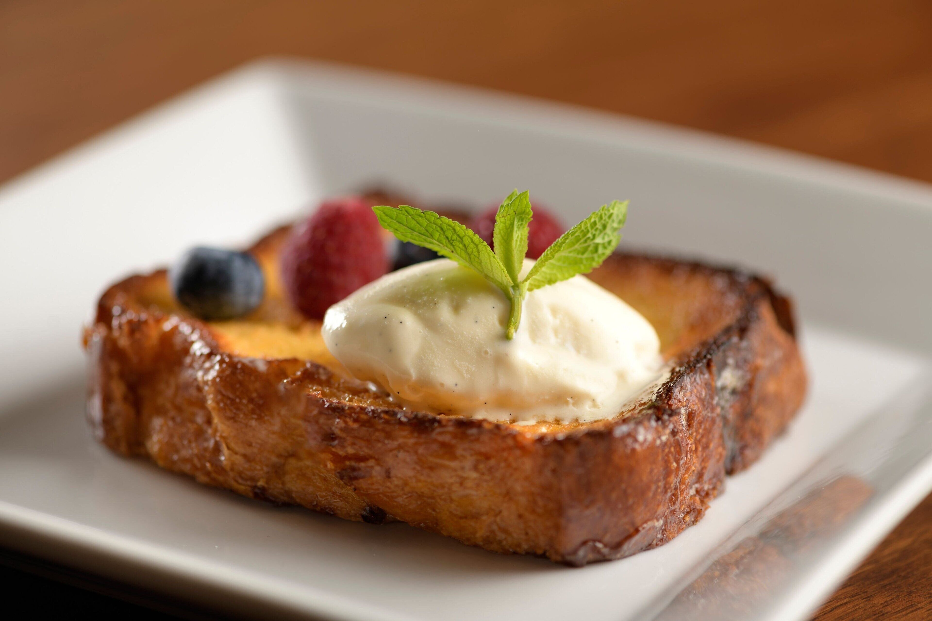 A plate with a slice of toast topped with a dollop of cream and a mint leaf, accompanied by berries.