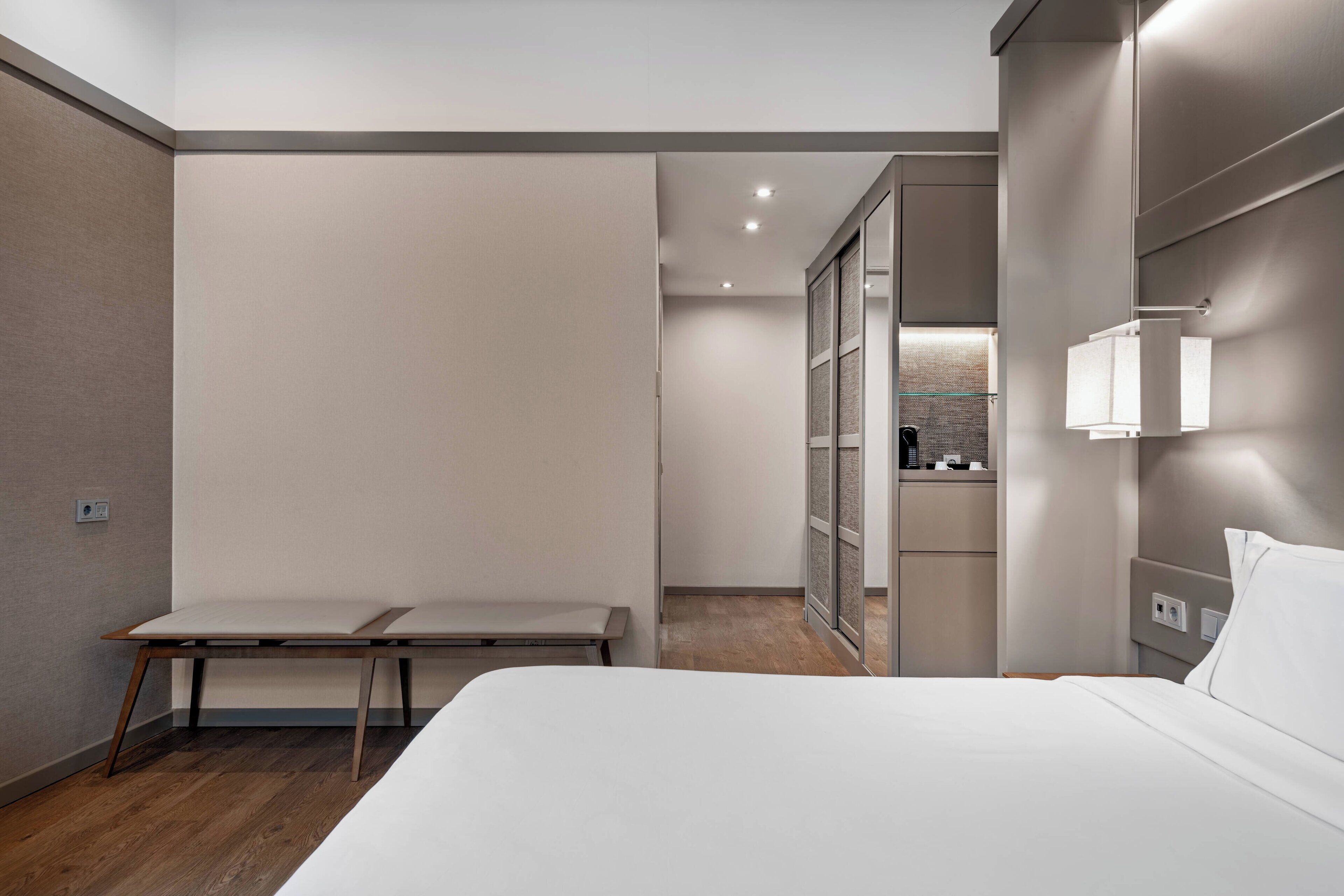 A modern hotel room with a bed, a bench, a desk, and a wardrobe.
