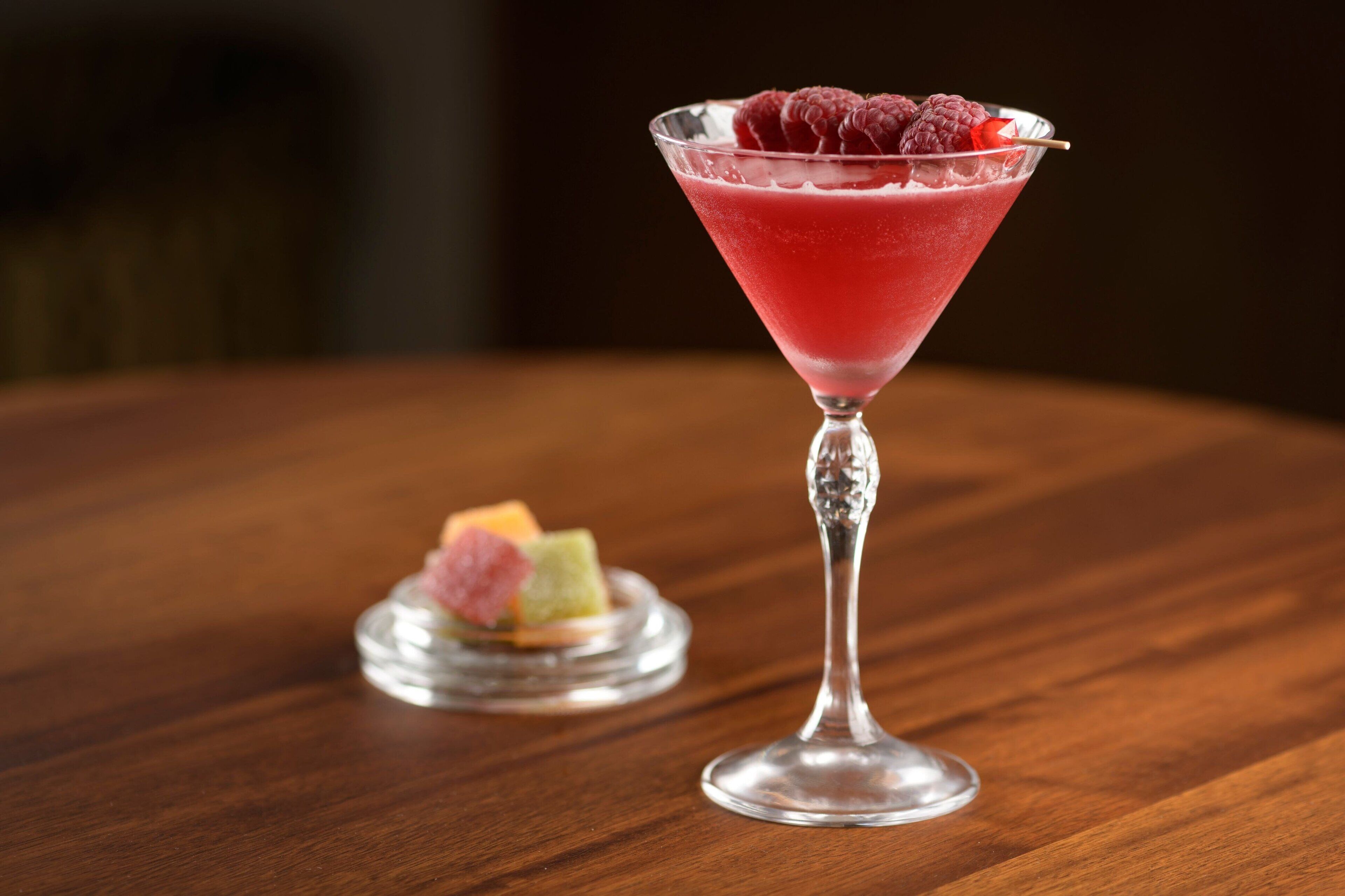 A martini glass with a red drink and raspberries on a wooden table.