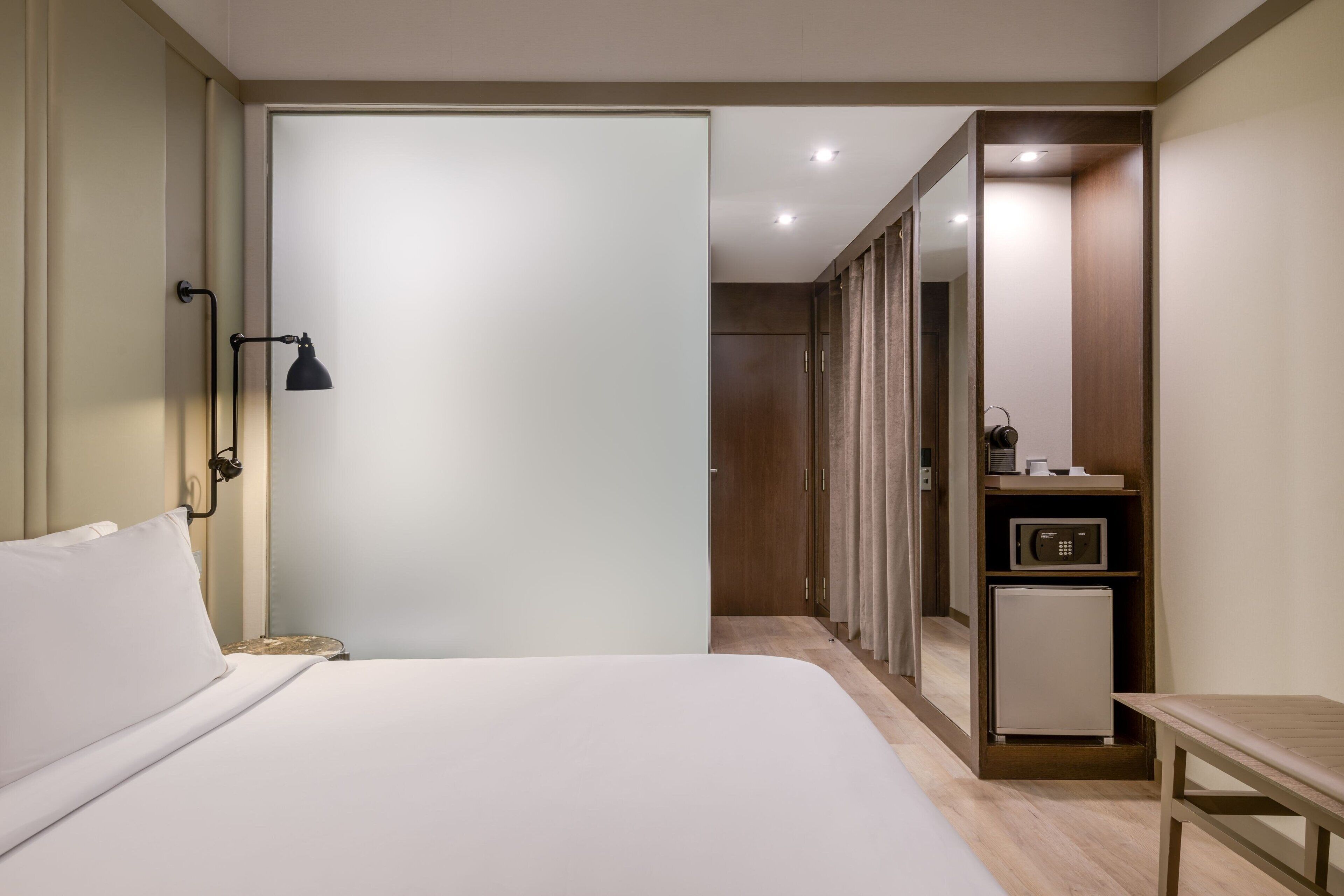 A hotel room with a bed, bedside lamp, a small table, a refrigerator, a mirror, and a wardrobe.