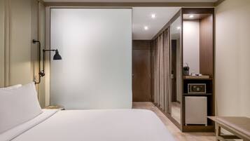A hotel room with a bed, bedside lamp, a small table, a refrigerator, a mirror, and a wardrobe.