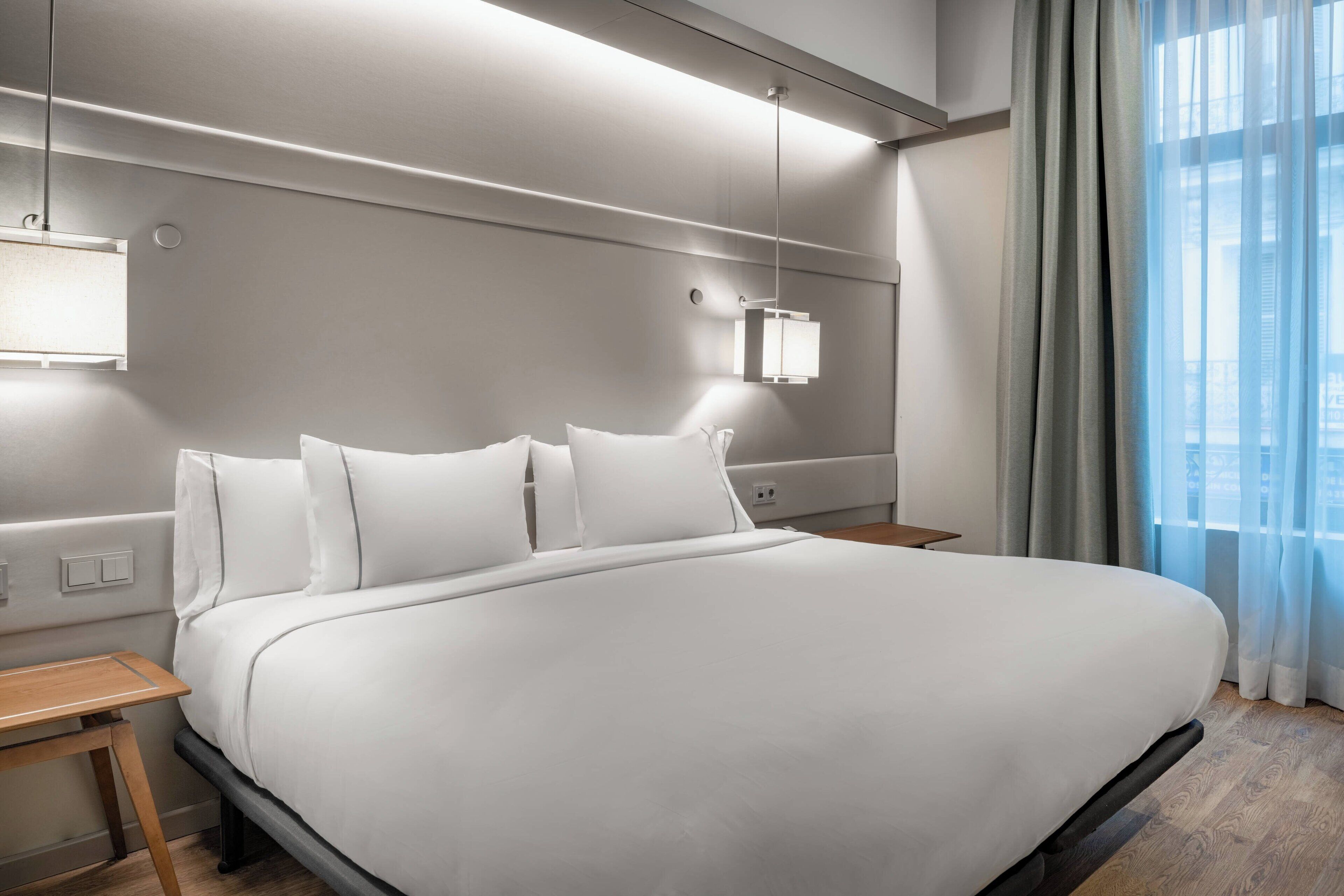 Una cama grande con ropa de cama blanca, una mesita de noche, una lámpara fijada a la pared y una ventana con cortinas.