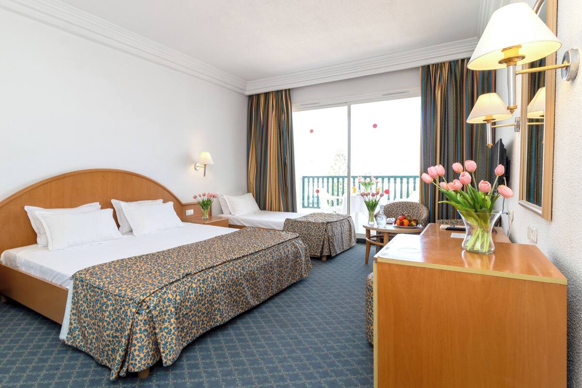 Une chambre d’hôtel avec un grand lit, un lit plus petit, un bureau et un balcon avec des rideaux.