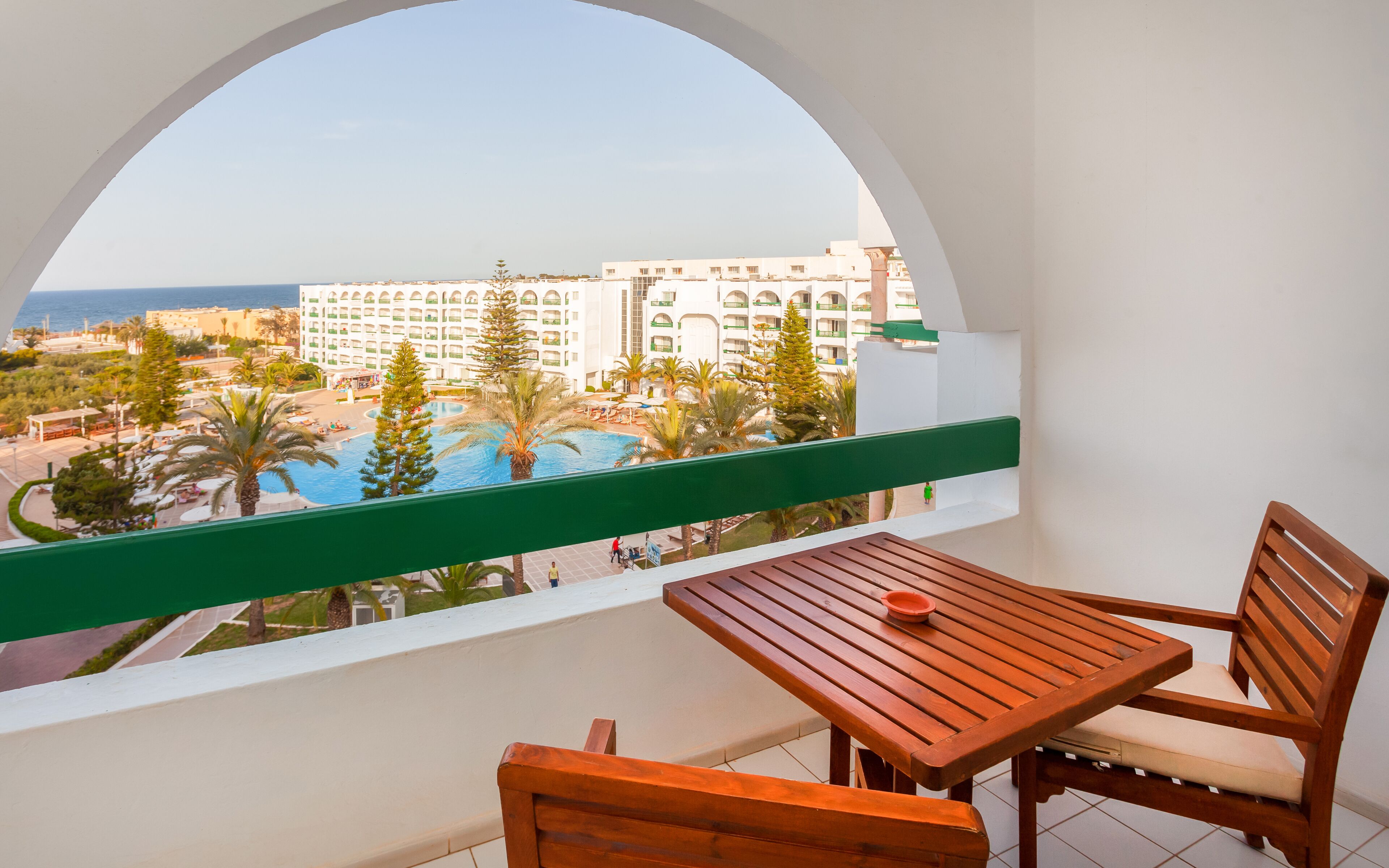 A balcony with a wooden table and chairs, overlooking a pool and a hotel building.