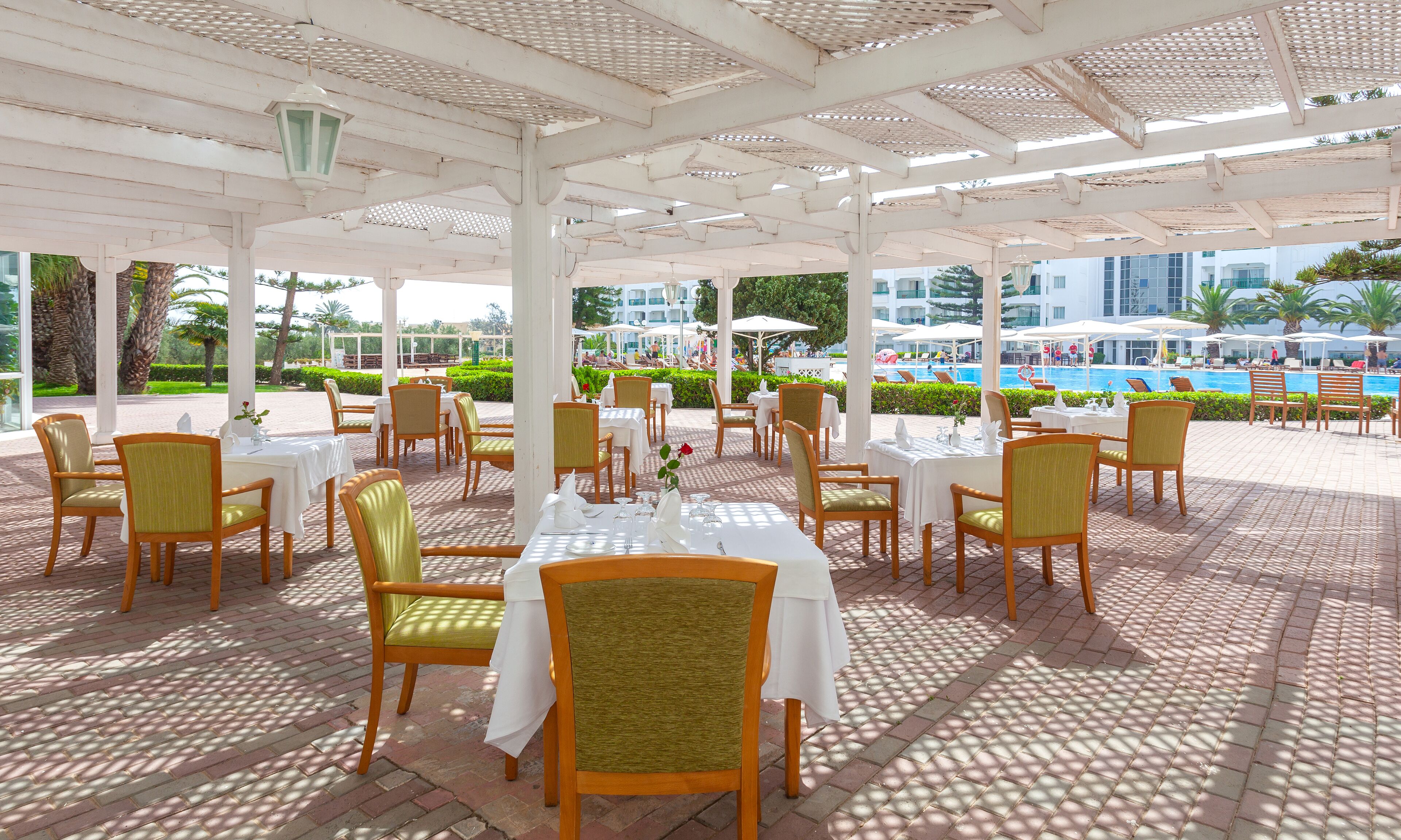 Un área de comedor al aire libre con mesas y sillas, una piscina al fondo y un edificio con banderas.