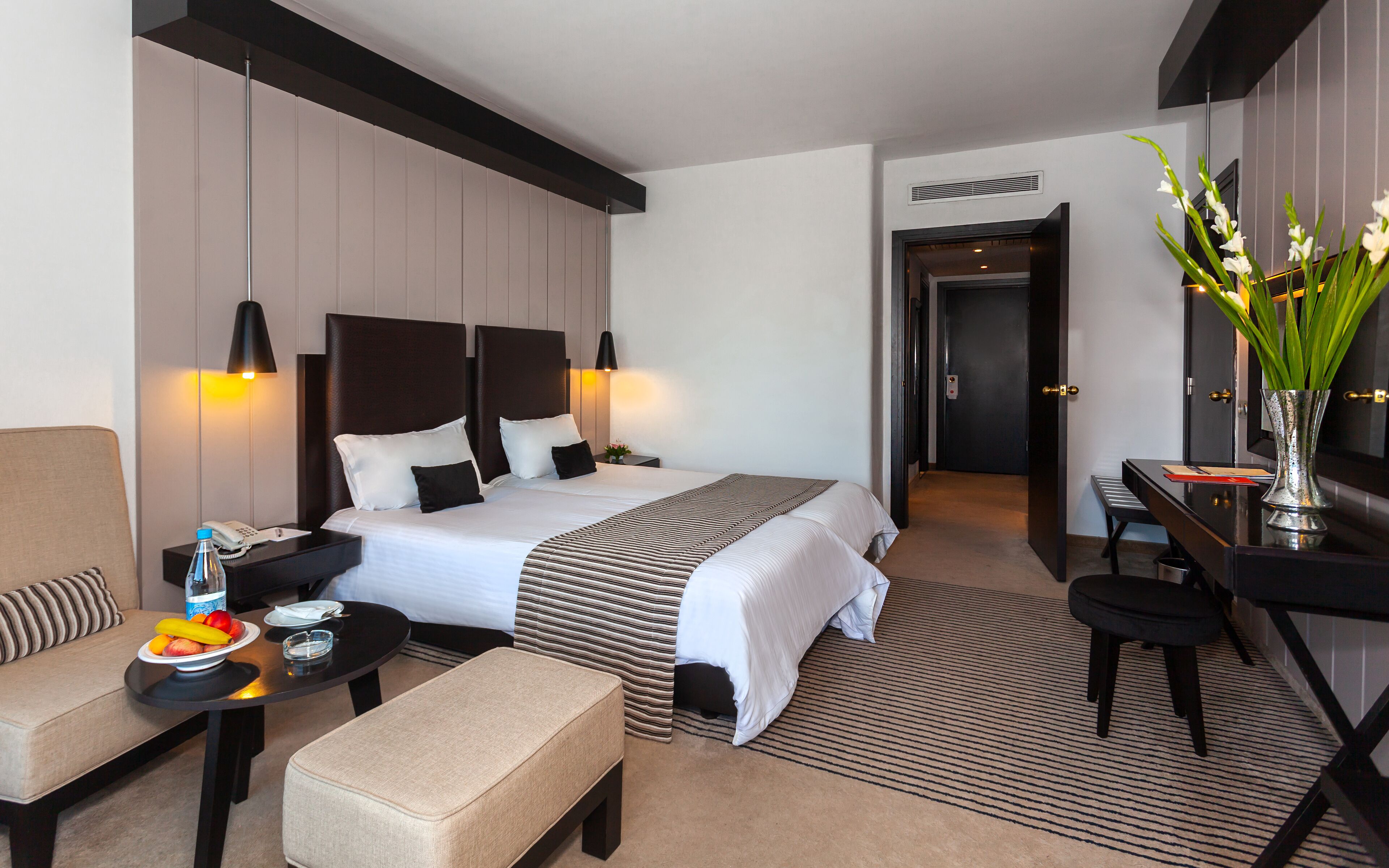 A hotel room with a large bed, bedside tables, a desk, a chair, a sofa, a small table with fruit, and a vase with flowers.