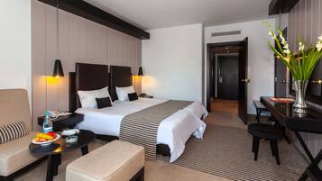 A hotel room with a large bed, bedside tables, a desk, a chair, a sofa, a small table with fruit, and a vase with flowers.