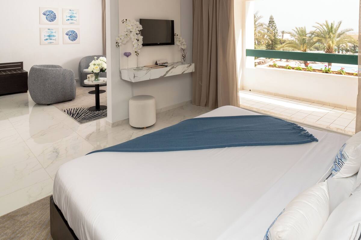 Une chambre d’hôtel moderne dotée d’un grand lit, d’un téléviseur fixé au mur, d’un petit coin salon et d’un balcon offrant une vue sur les palmiers.