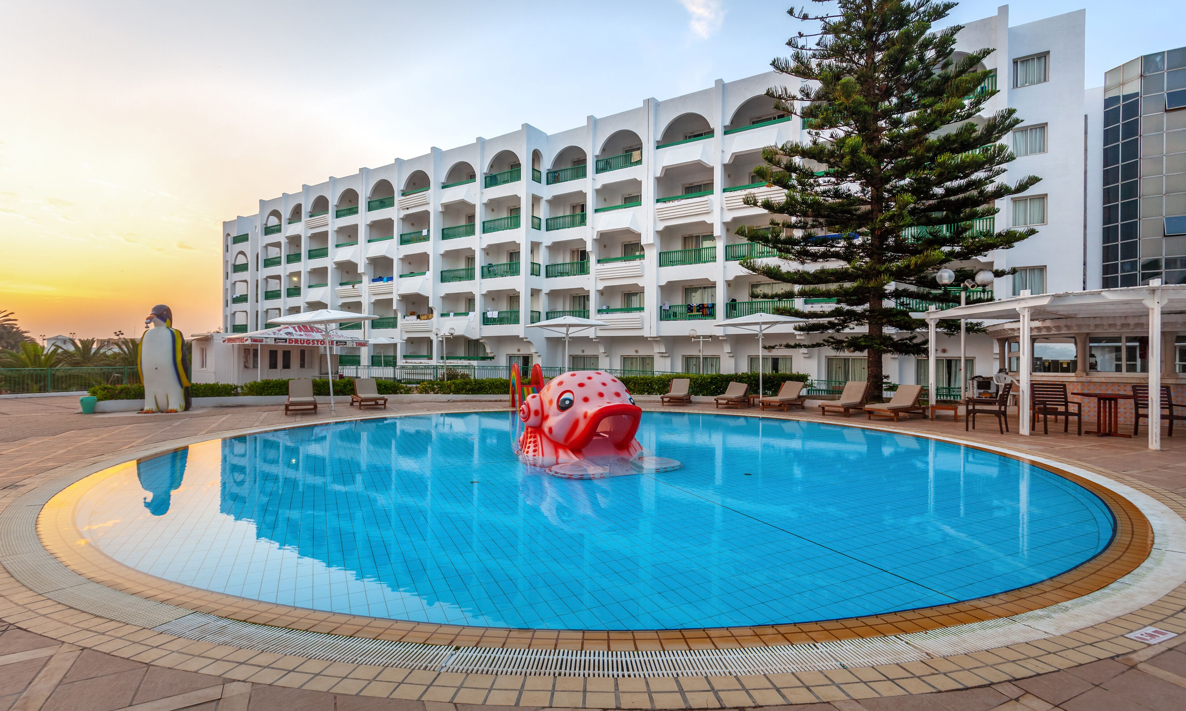 Un hôtel doté d’une piscine, un grand bâtiment avec de nombreux balcons et un arbre tropical.