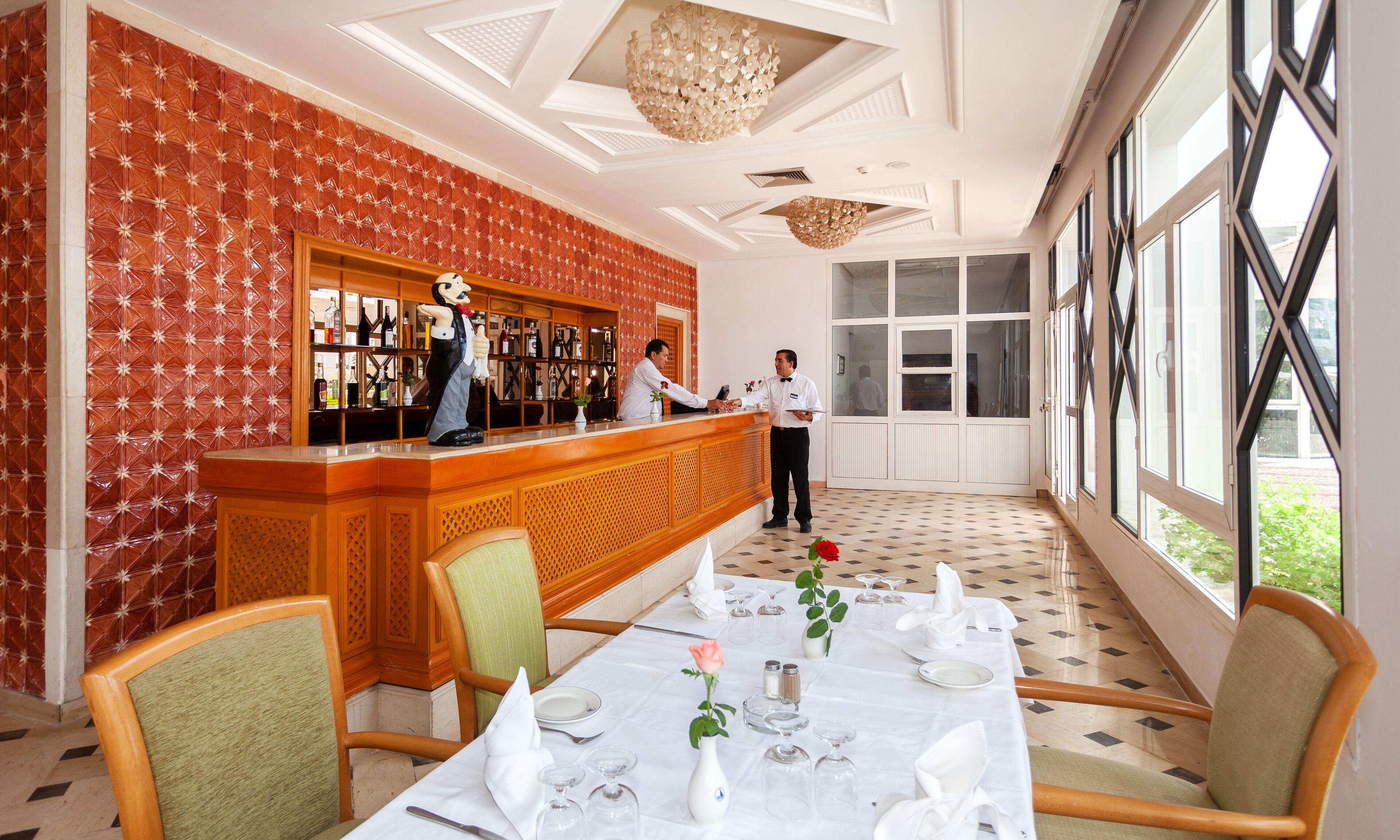 A hotel lobby with a reception desk, a bar area, and a dining area with tables set for guests.