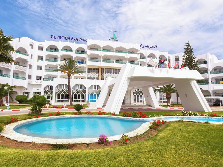 Un hotel con piscina, palme e un grande edificio bianco dotato di numerosi balconi e bandiere.