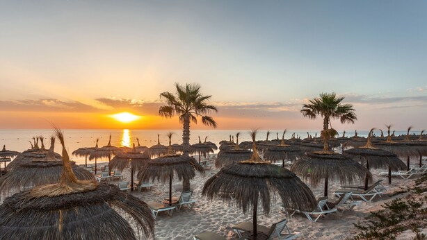 Una playa con sombrillas de paja y sillones reclinables, un atardecer de fondo y palmeras.