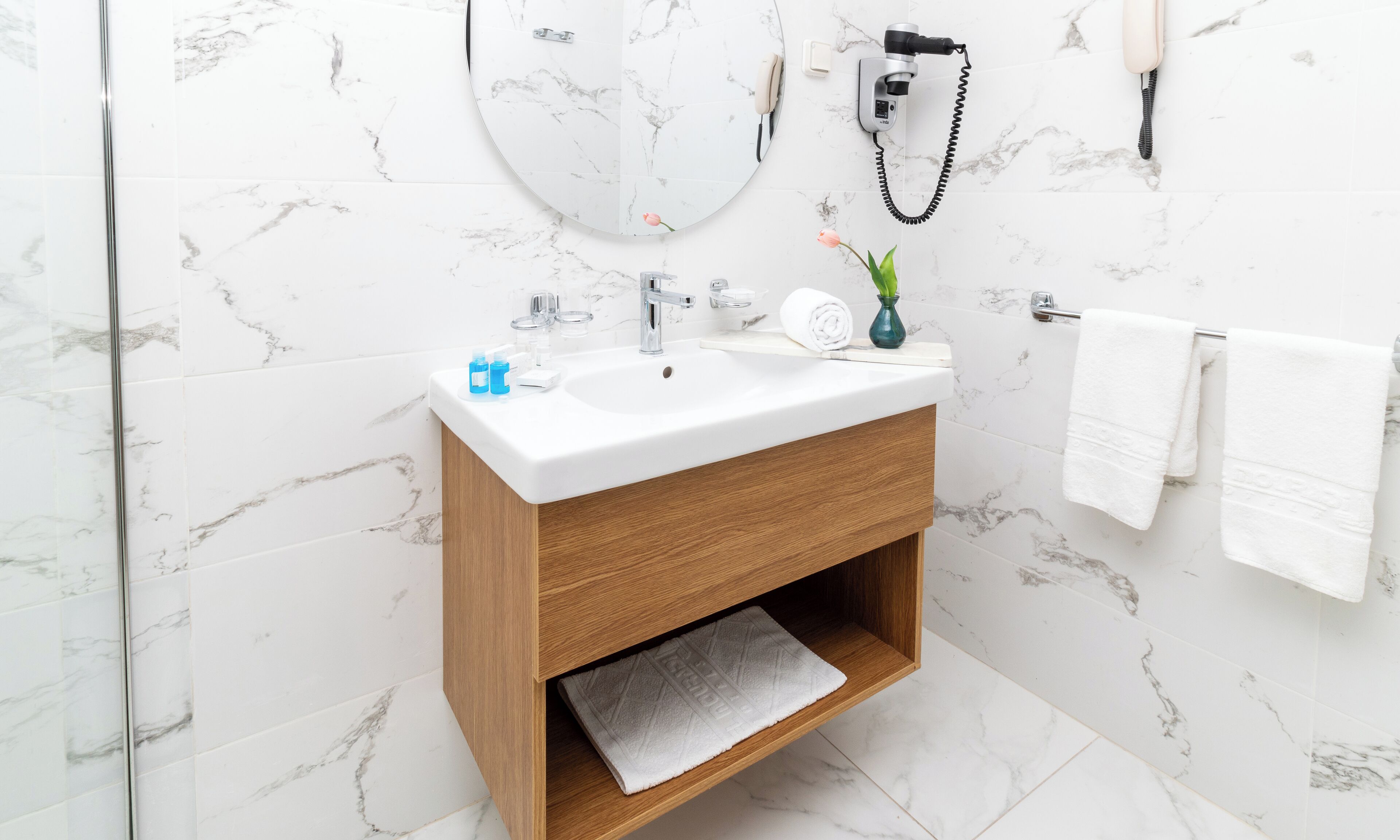 Ein Badezimmer mit marmorverkleideter Wand, einem weißen Waschbecken mit Holzschrank, einem Handtuchhalter und einem Fön an der Wand.