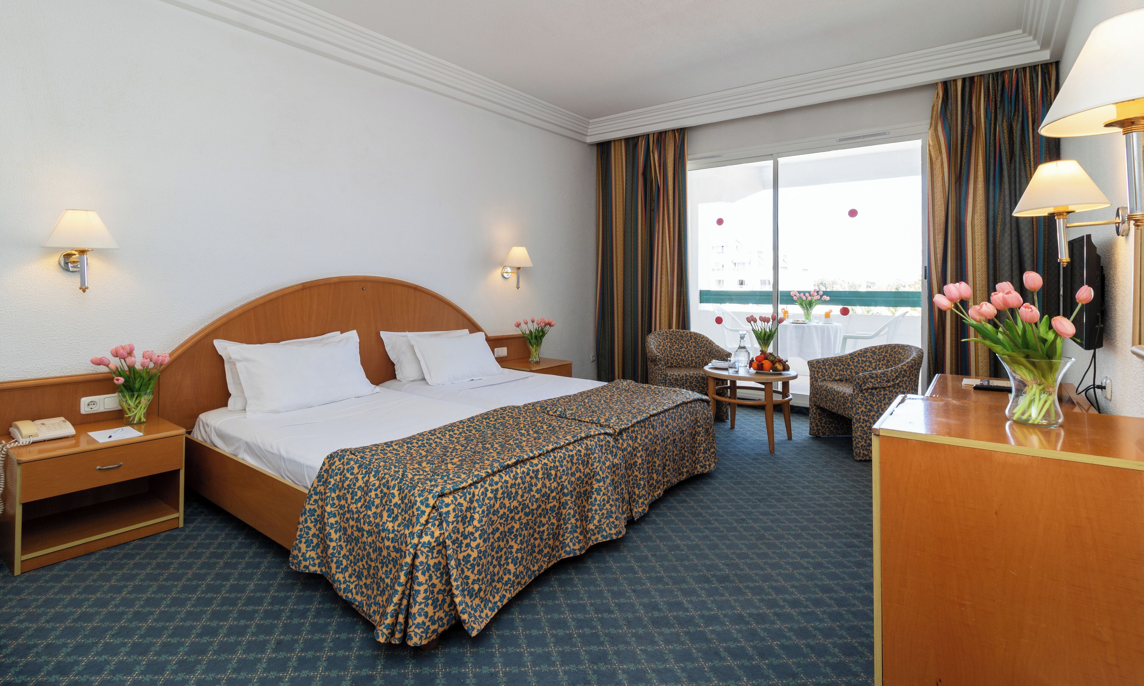 Een hotelkamer met een groot bed, twee nachtkastjes, een bureau met stoel, een klein tafeltje met een bloevas en uitzicht op zee.