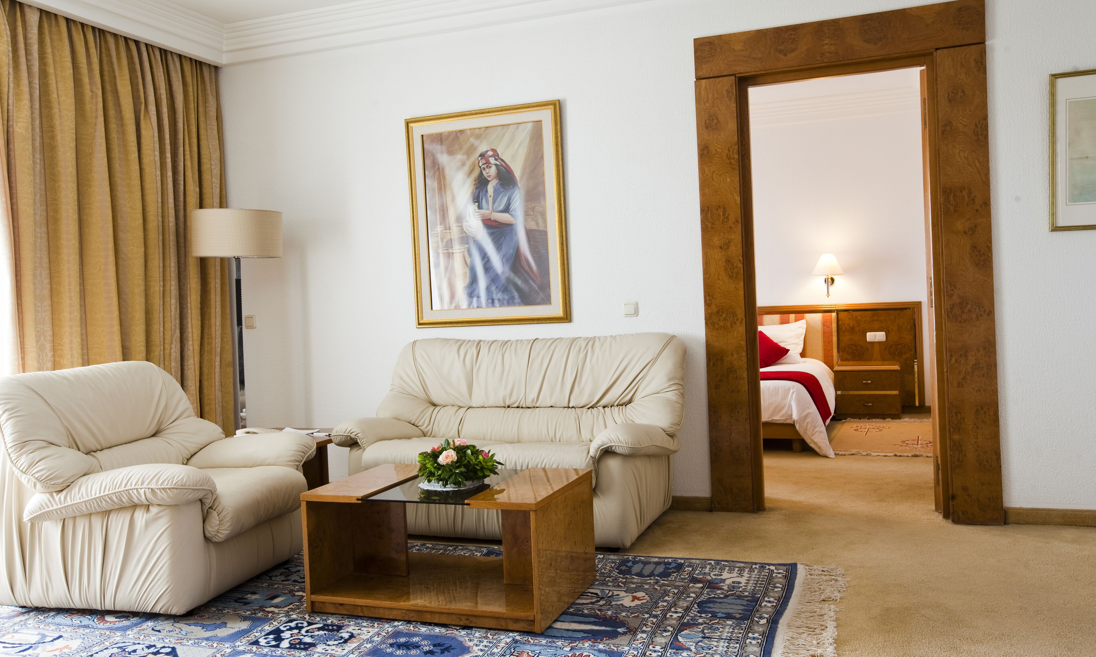 A hotel room with a beige sofa, a wooden coffee table, a patterned rug, a painting on the wall, and a wooden door leading to another room.