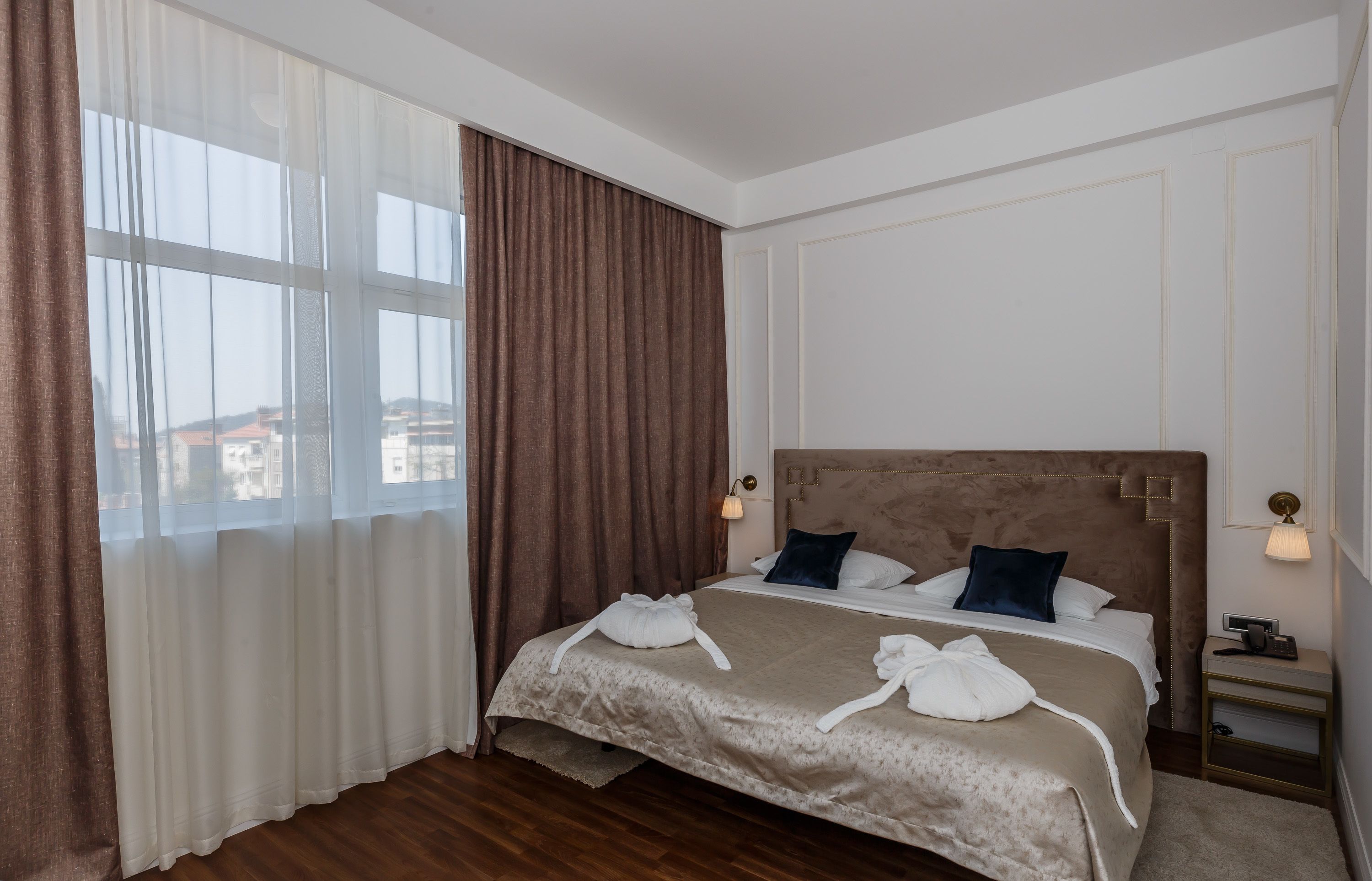 A hotel room with a large bed, a nightstand, a lamp, and a view of buildings through the window.