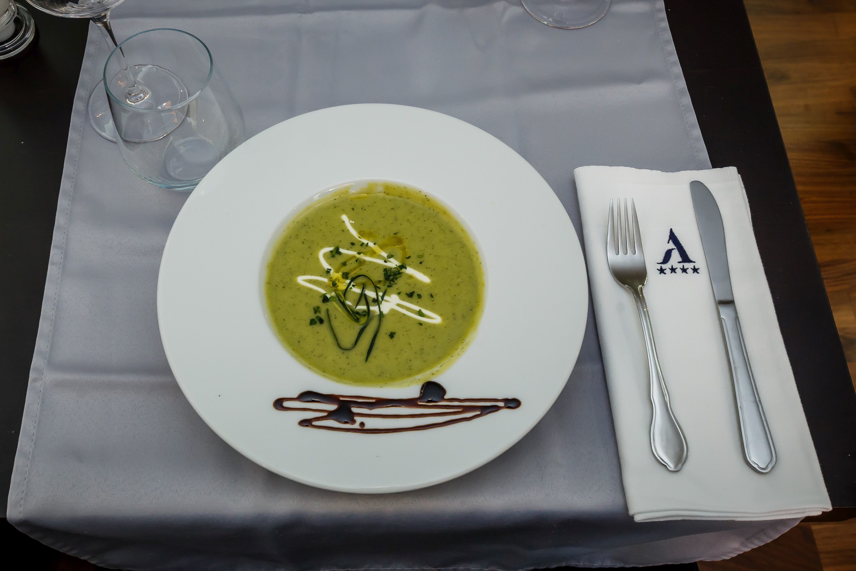 Un plato blanco con sopa verde adornada con hierbas y salsa, acompañado de un servilleta doblada y cubiertos sobre un mantel.