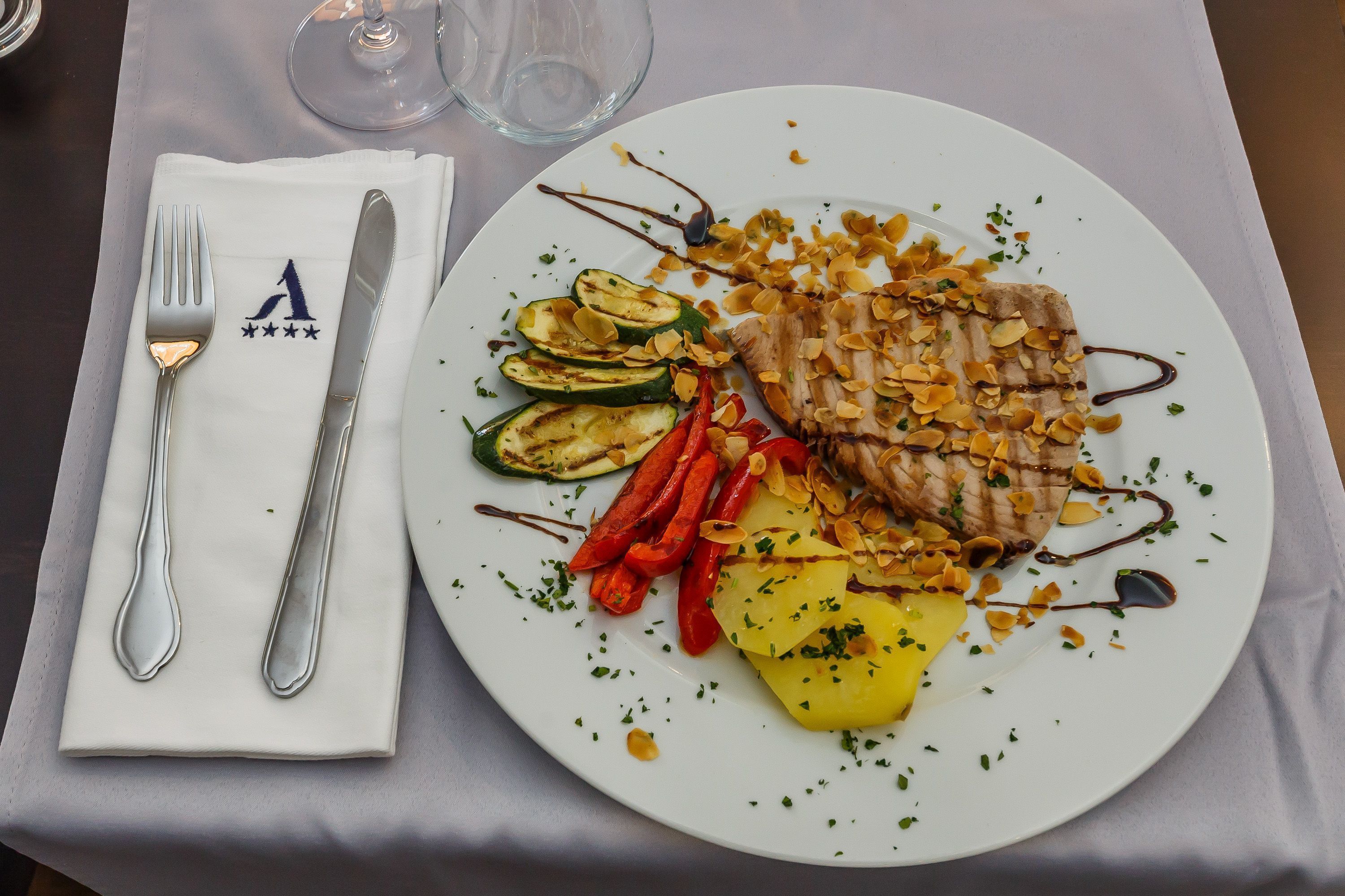 A plate of food with grilled meat, zucchini, red peppers, and pineapple, garnished with herbs and nuts, on a white plate.