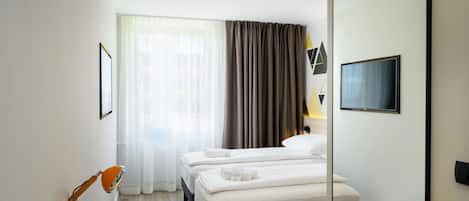A hotel room with a bed, a desk with a chair, a TV mounted on the wall, and a window with curtains.