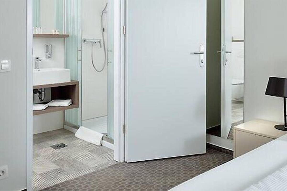 A hotel room with a bed, a bedside table with a lamp, a bathroom with a shower, and a sink.