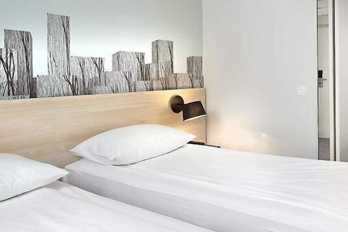 A hotel room with two beds, a wooden headboard, a wall-mounted lamp, a desk with a chair, and a door leading to another room.