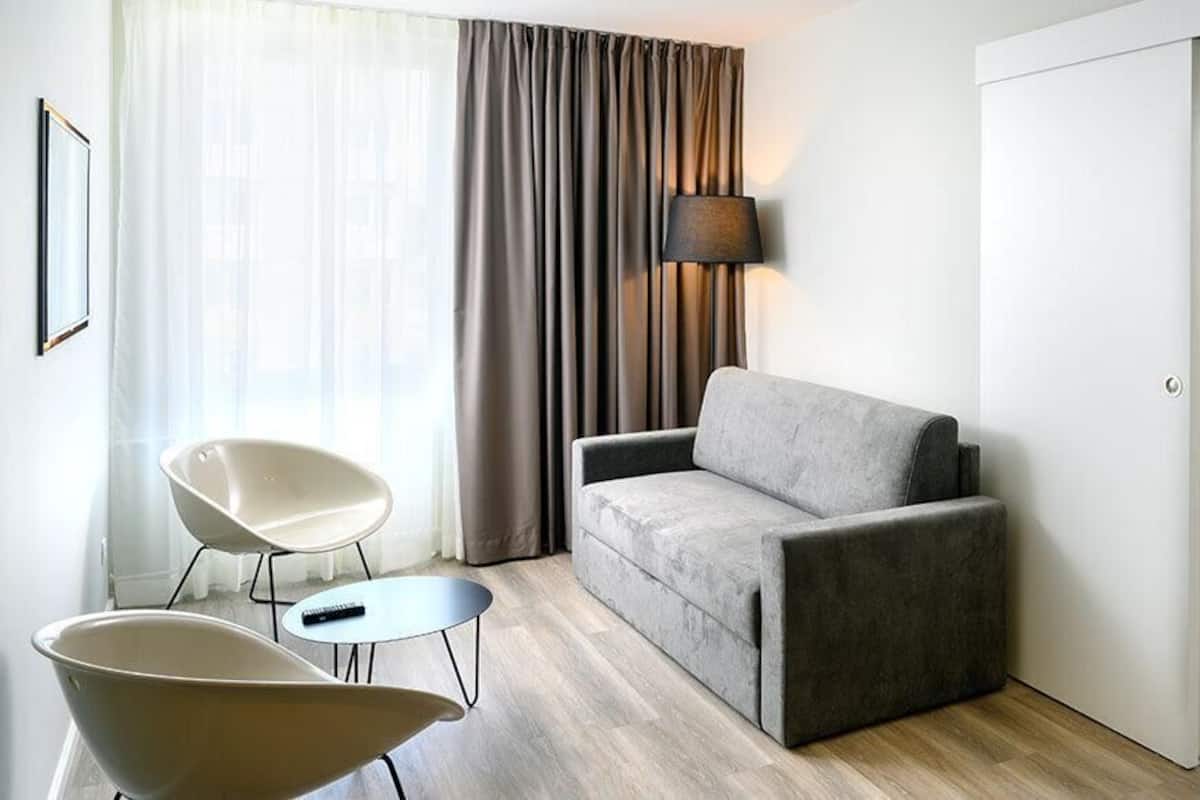 A modern hotel room with a grey sofa, a white armchair, a small round table, and a bed with a white duvet.