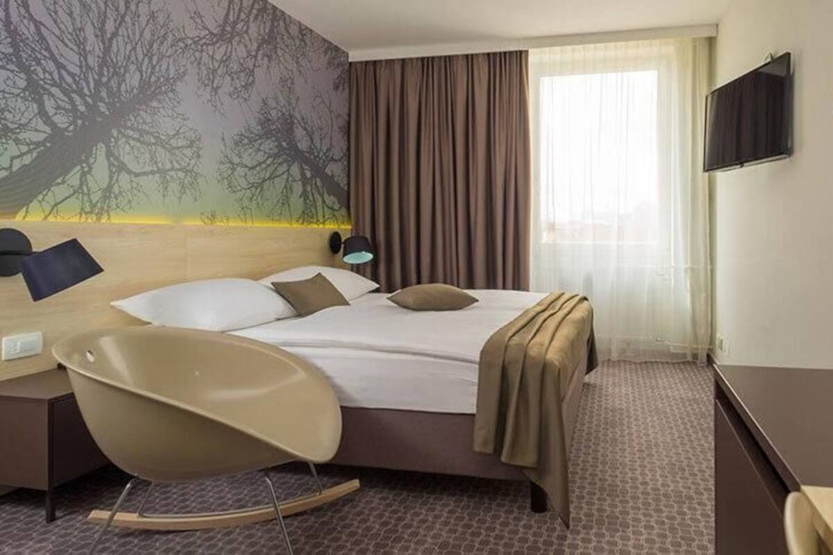 A hotel room with a bed, a chair, a desk, a lamp, a TV, and a wall with a tree mural.