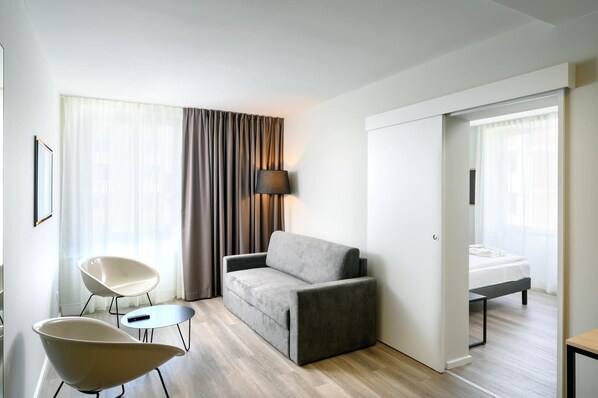 Una habitación de hotel moderna con un sofá gris, dos sillones, una mesita y una cama con un banco.