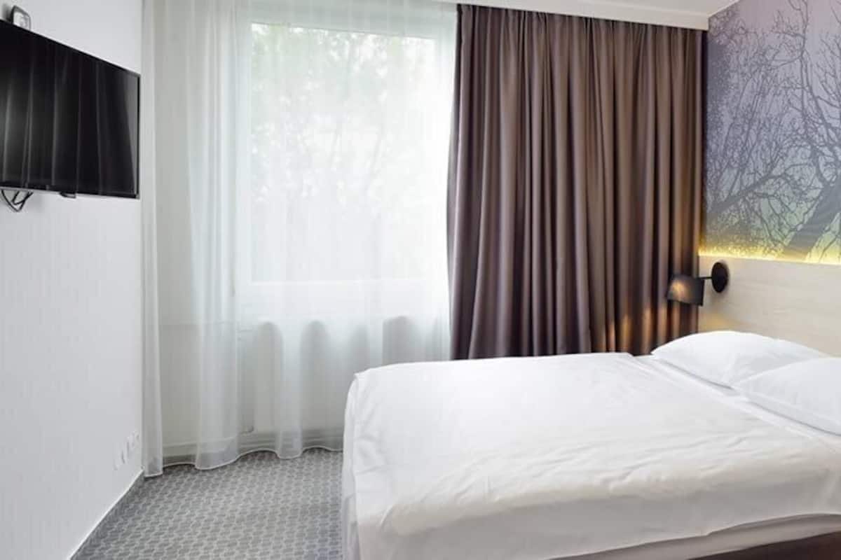 A hotel room with a bed, a nightstand, a wall-mounted TV, and a window with curtains.