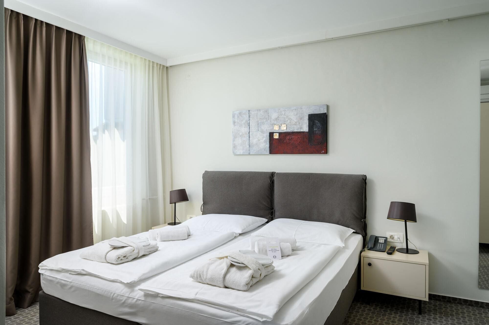 A hotel room with a bed, two bedside tables, a painting on the wall, and a window with curtains.