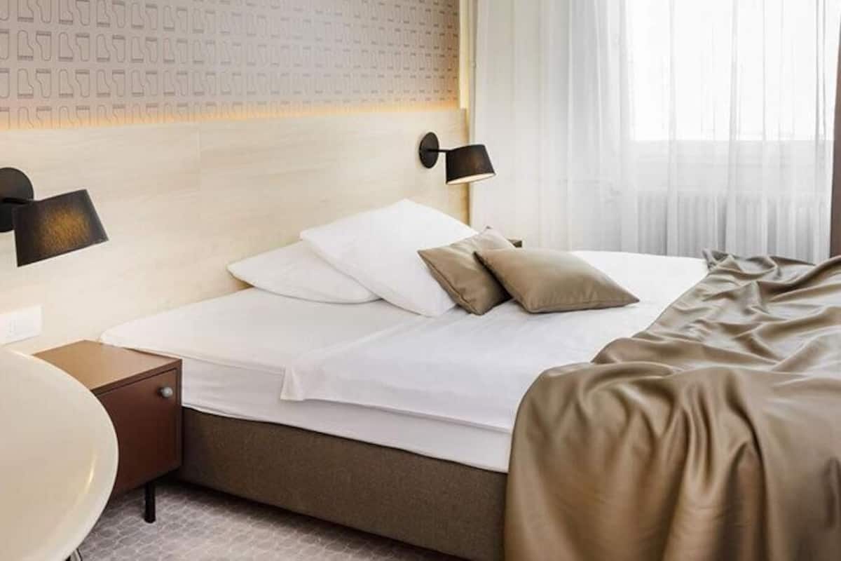 A modern hotel room with a large bed, a bedside table, a wall-mounted lamp, and a large bathtub.