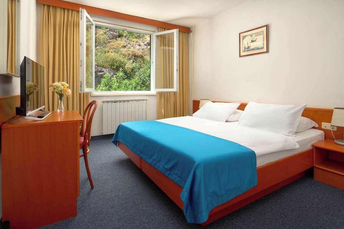 Quarto de hotel com cama, escrivaninha com cadeira, janela com cortinas e radiador.