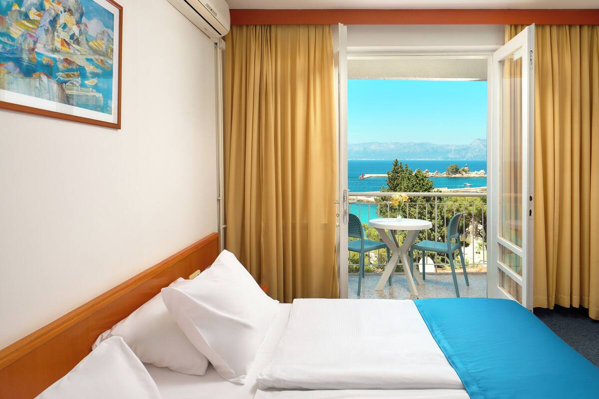 Quarto de hotel com uma cama, um quadro na parede e uma varanda com mesa e cadeiras.