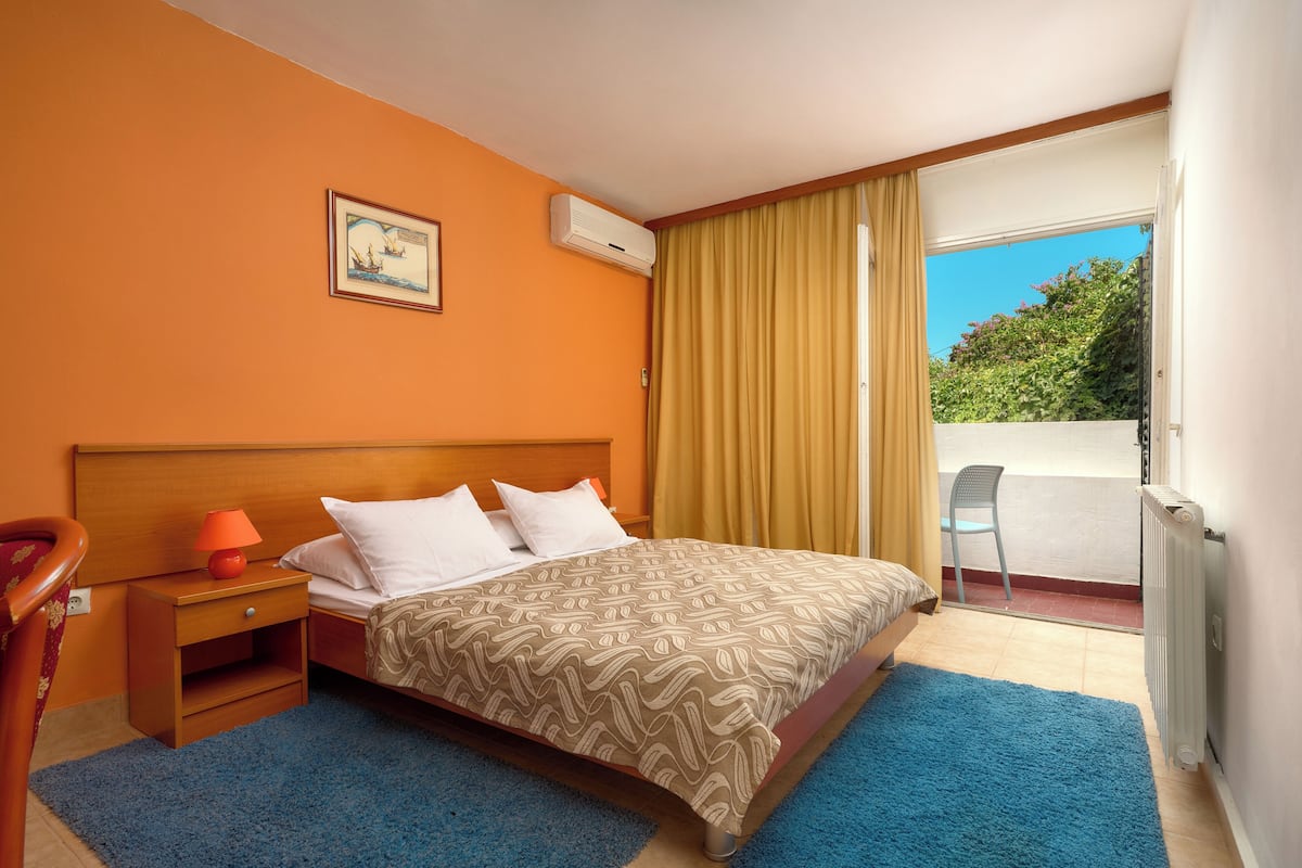 Quarto de hotel com cama, mesa de cabeceira, cadeira, janela com cortinas e vista para o verde lá fora.