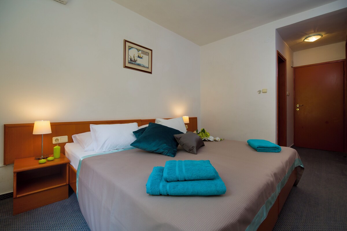 Una habitación de hotel con una cama, dos mesitas de noche, una lámpara y un cuadro en la pared, además de una puerta.