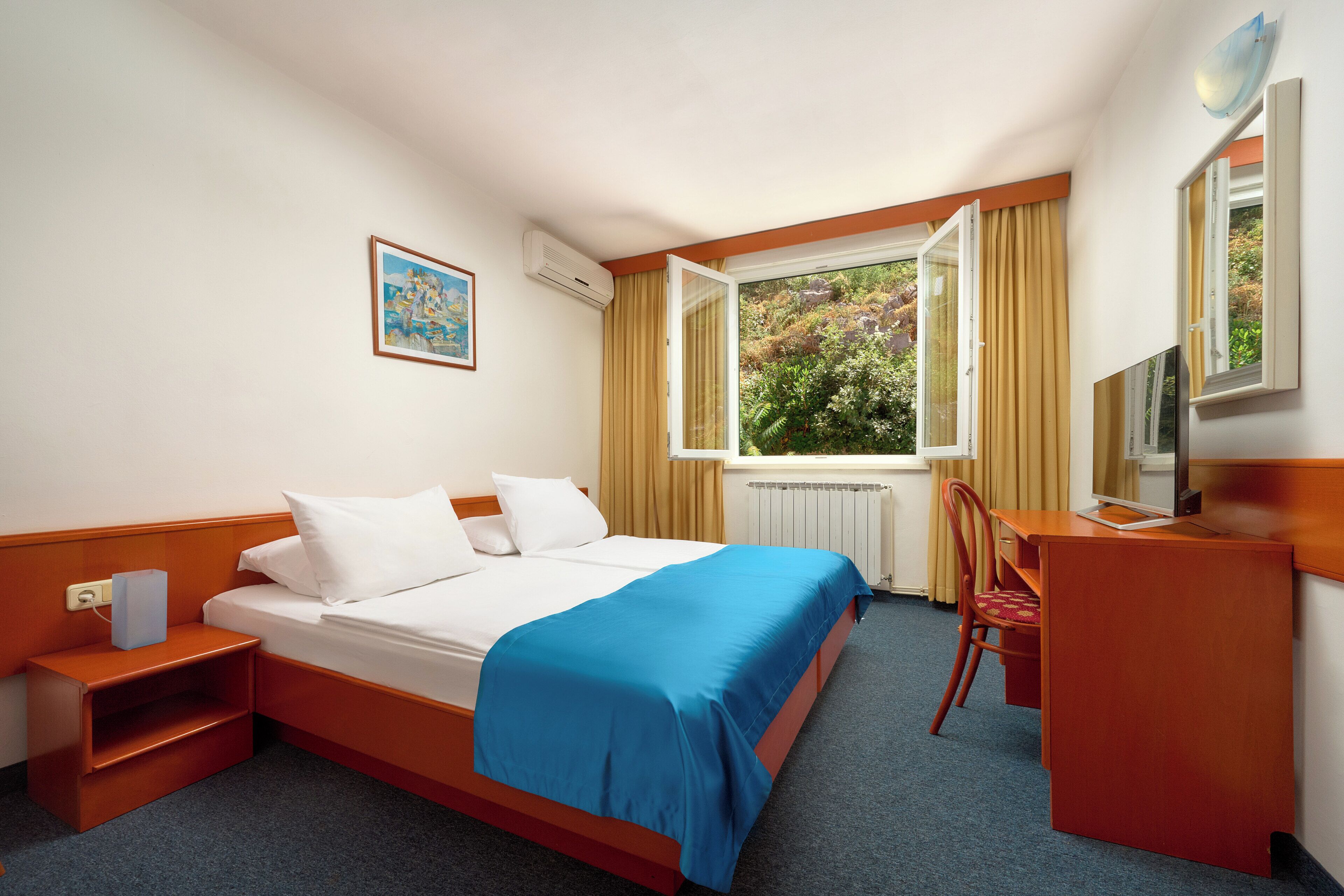 Ein Hotelzimmer mit Bett, Schreibtisch, Stuhl, einem Fenster mit Vorhängen, einem Heizkörper und einem Bild an der Wand.