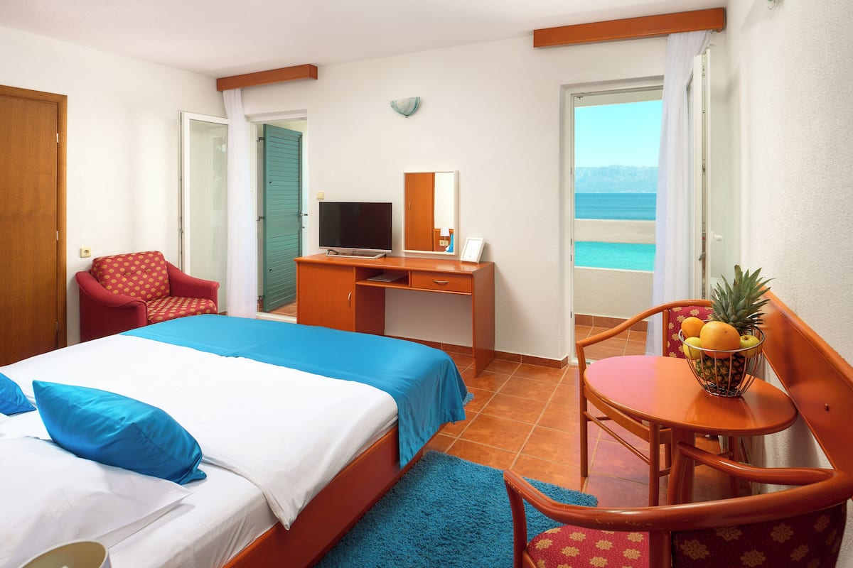 Quarto de hotel com uma cama, uma escrivaninha com televisão, uma cadeira, uma mesinha com fruta e vista para o mar.