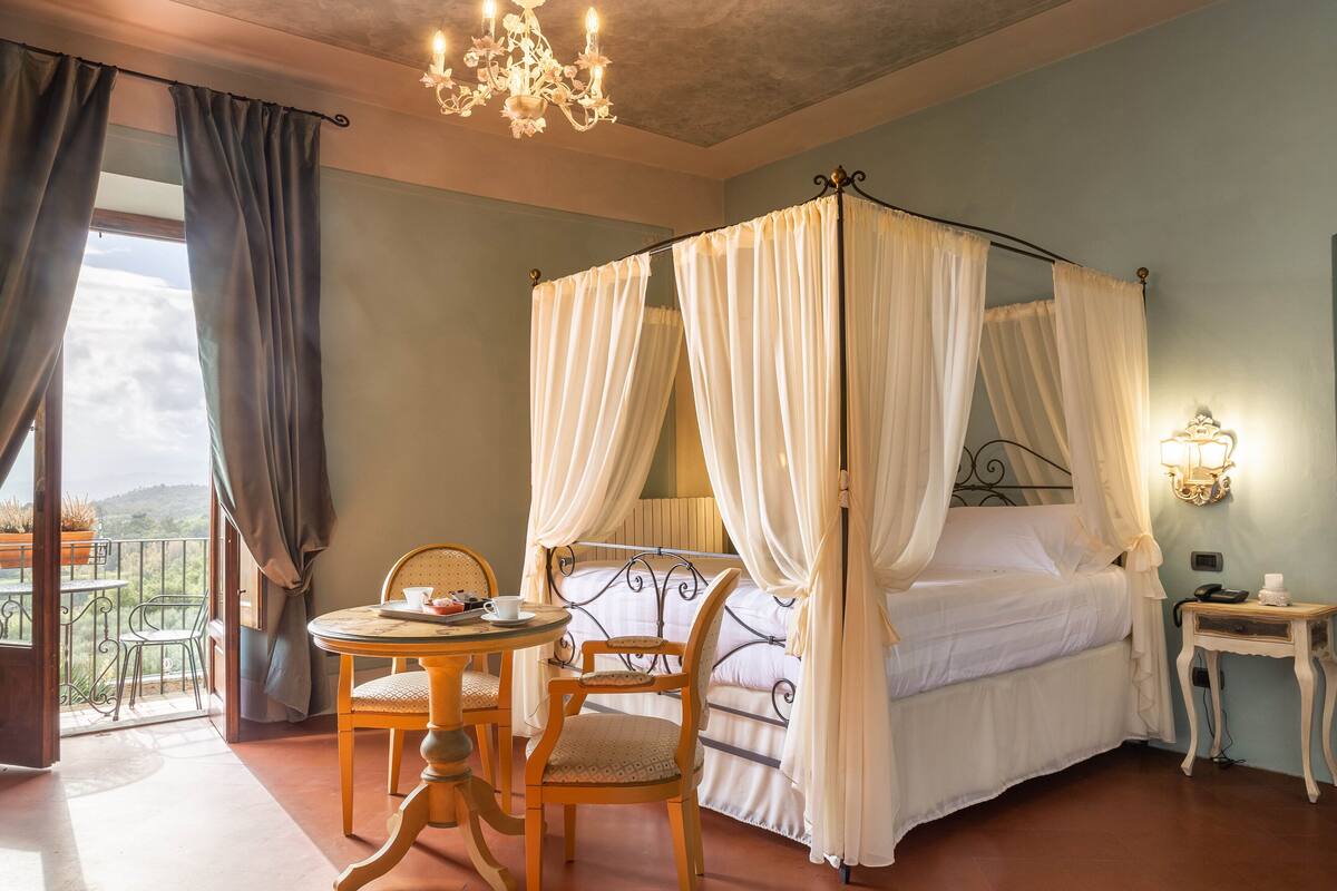 Un lit à baldaquin avec du linge de lit blanc, une table ronde en bois avec des chaises, une petite table de chevet avec une lampe, et un balcon offrant une vue sur les collines.