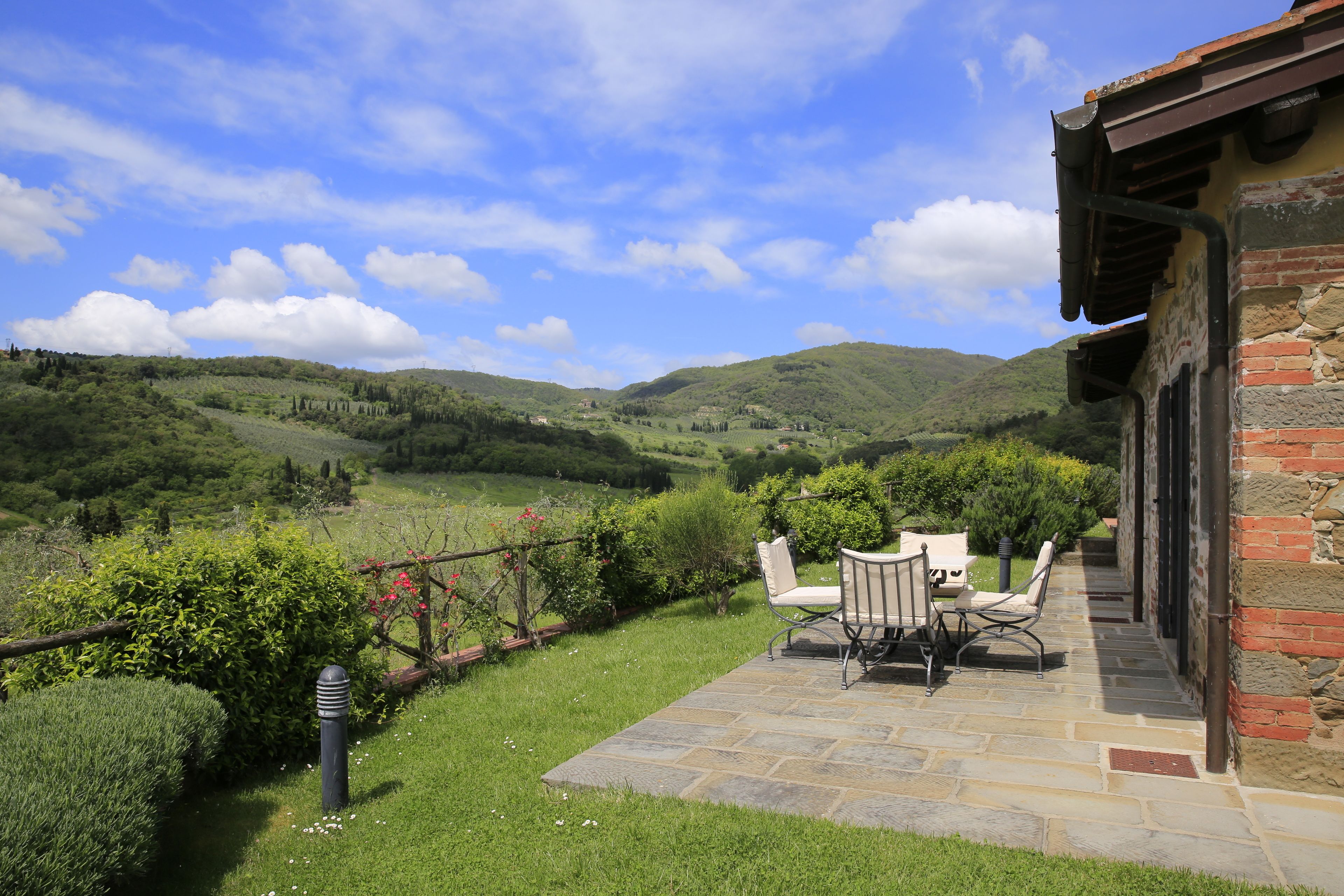 A patio with stone flooring and two chairs, overlooking a lush valley with hills and trees.