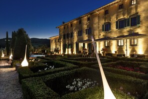 Un jardin bien entretenu, agrémenté de haies géométriques et d’un chemin menant à un bâtiment doté de nombreuses fenêtres et d’un espace extérieur aménagé pour s’asseoir.