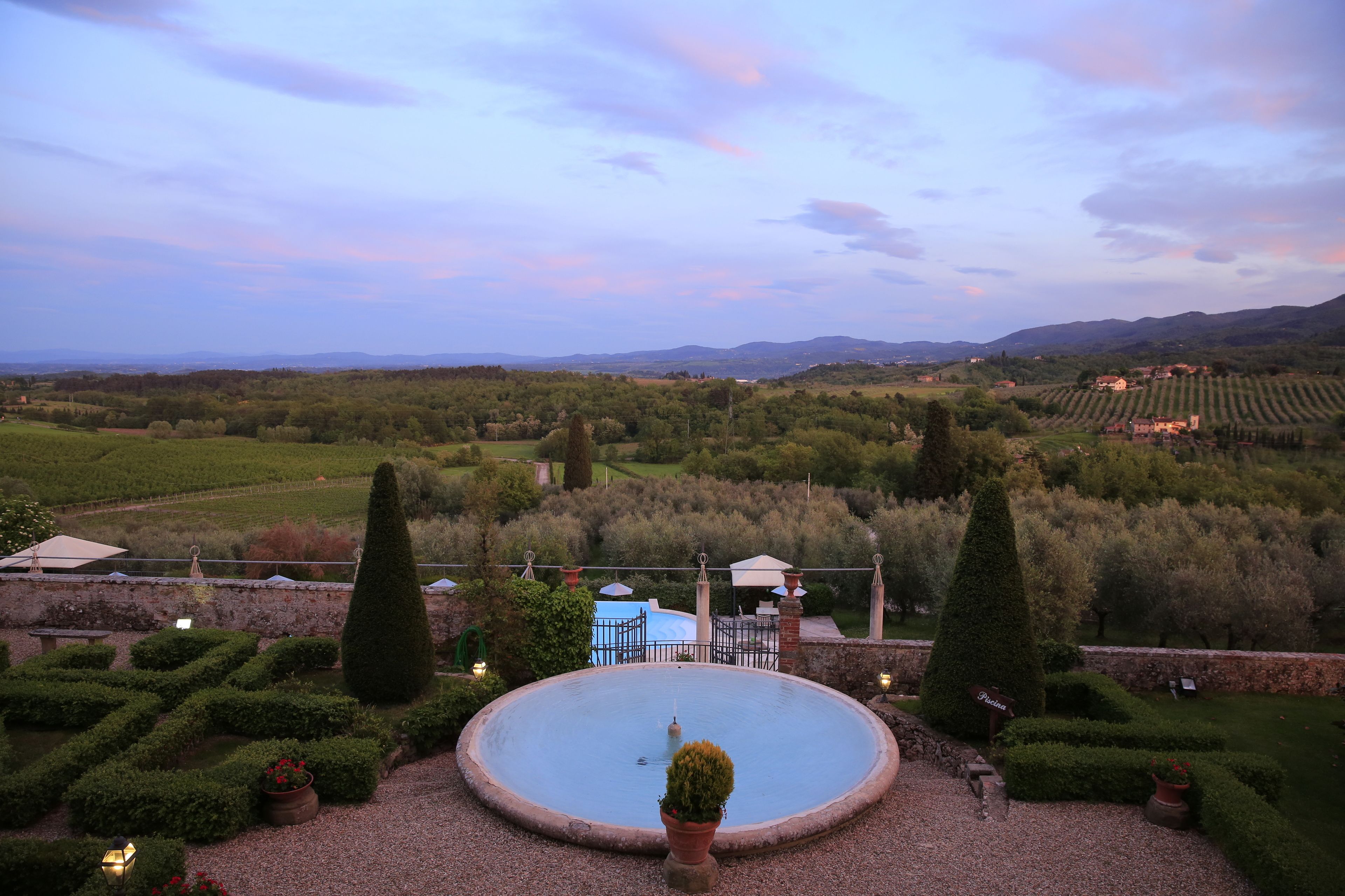 A circular pool surrounded by neatly trimmed hedges and potted plants, with a view of vineyards and distant hills in the background.