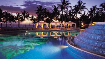 Una piscina con agua azul cristalina, rodeada de sillas de descanso y palmeras, con un atardecer de fondo.